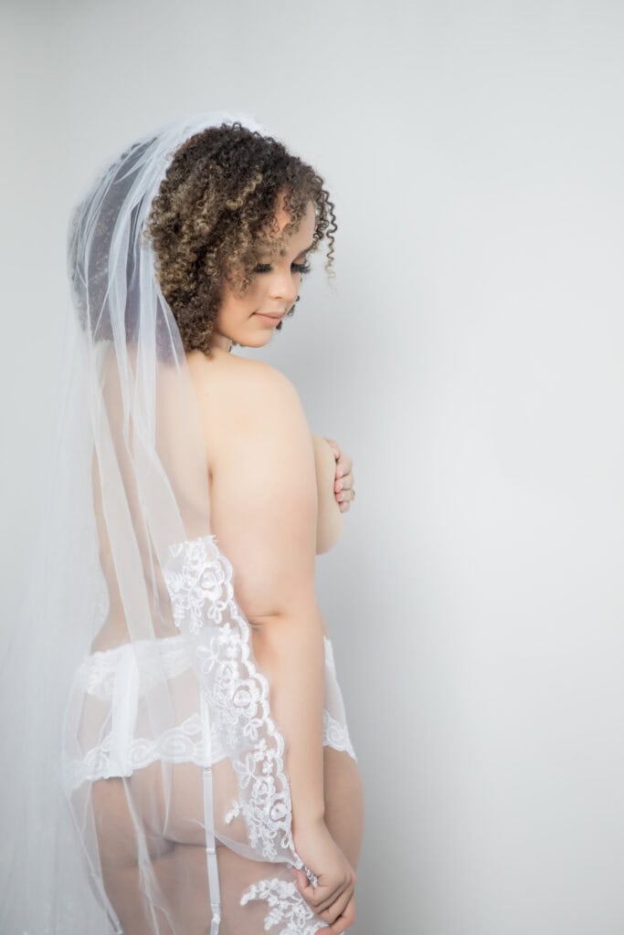 A pregnant bride with curly hair wearing a lace gown and veil, looking over her shoulder against a grey background.