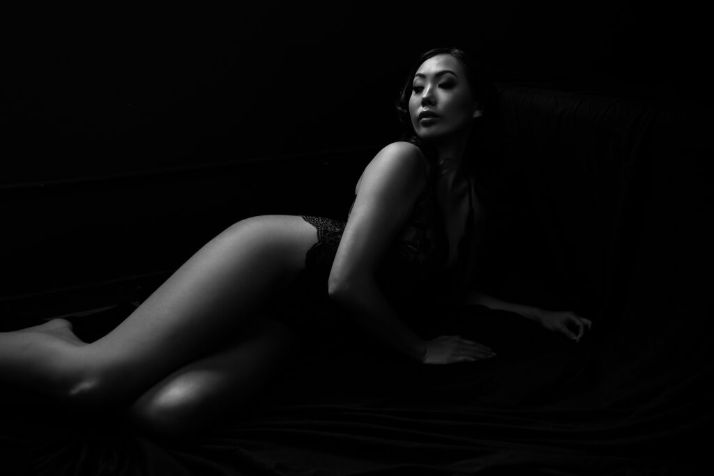 A black and white artistic portrait of a woman lying on a dark cloth, dressed in a lace outfit, looking contemplatively to the side.