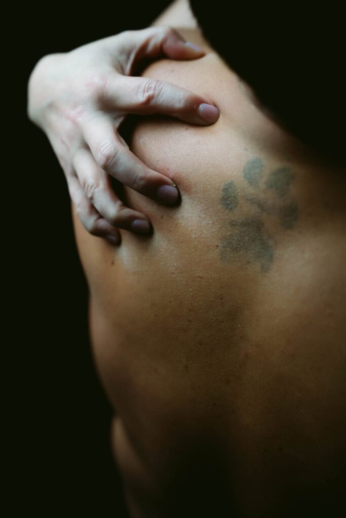 A person's hand touching their shoulder with a faded tattoo, set against a dark background.