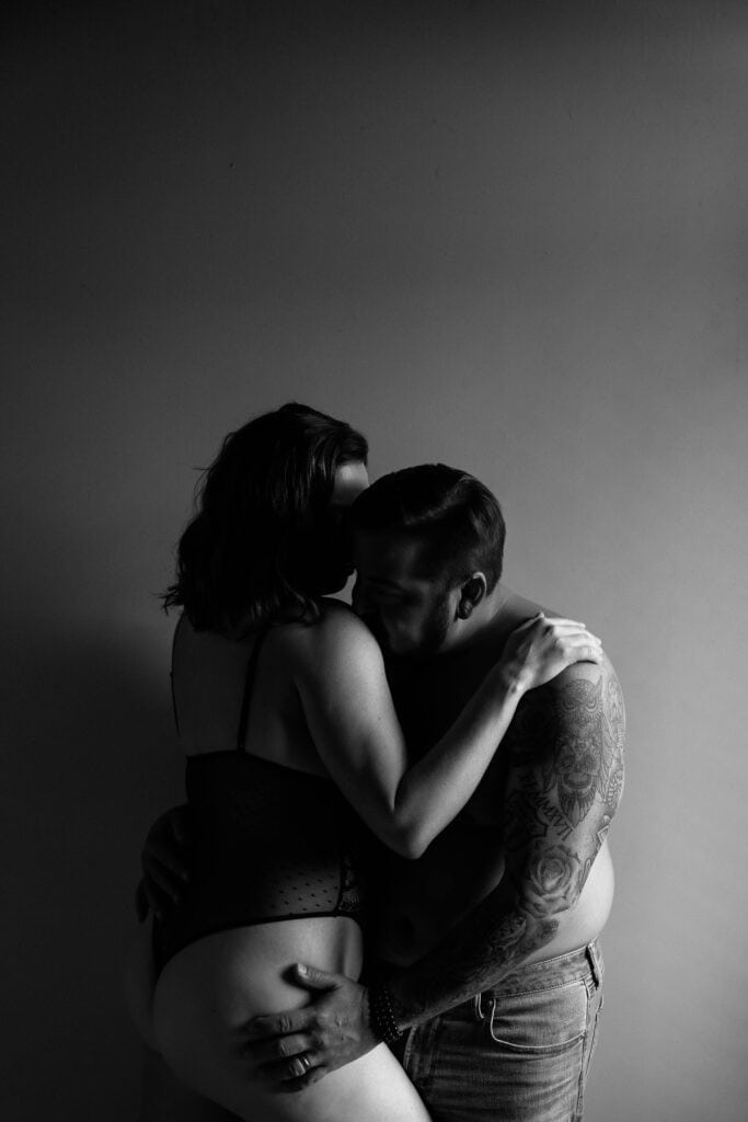 A black and white photo of a tattooed man and a woman embracing and kissing intimately against a plain background.