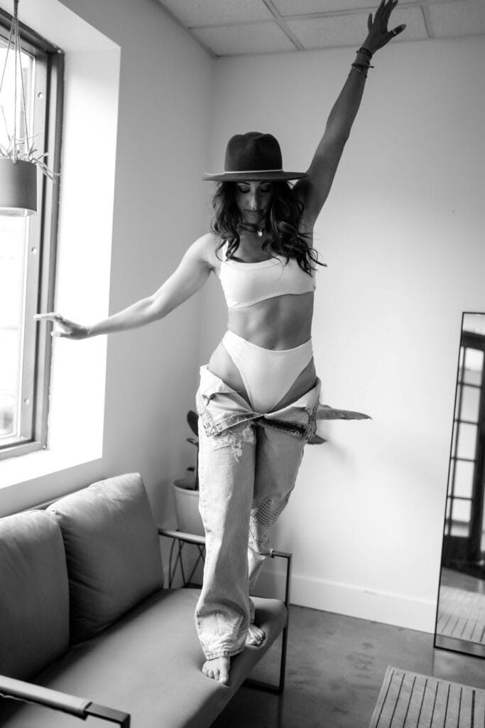 A woman in a white crop top and jeans with a tan hat dances on a sofa in a room with natural light.