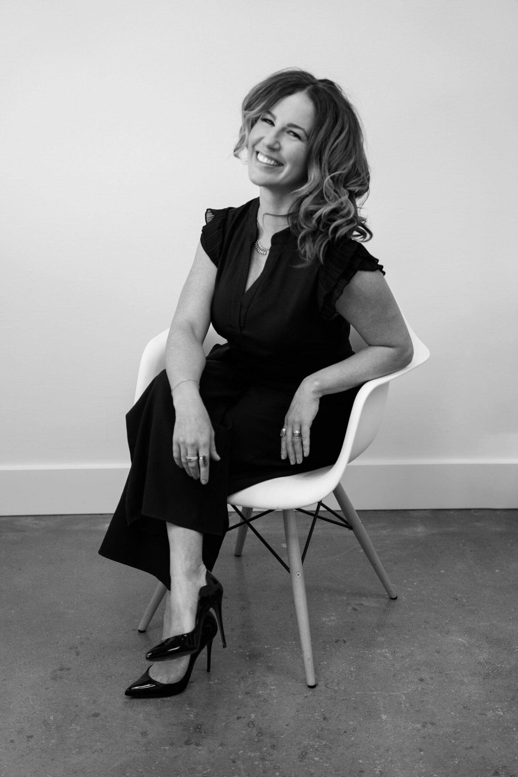 Black-and-white photo of a woman sitting on a modern chair, smiling. She has shoulder-length wavy hair and is wearing a dark short-sleeved outfit with ruffles, paired with high heels. The floor is concrete, and the background is a plain white wall. Image by Peekaboo Boudoir studio at Portland and Hood River.